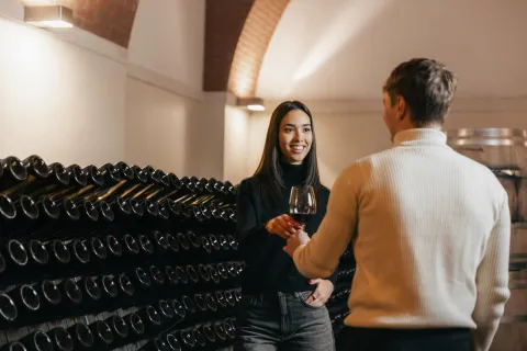 Un ragazzo e una ragazza si passano un calice di vino rosso in una cantina con bottiglie sullo sfondo