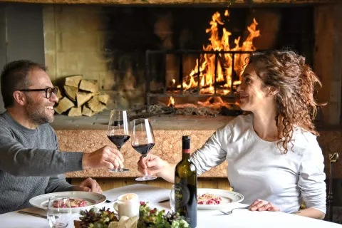 Due presone a cena in malga brindano con del vino rosso e con il fuoco acceso