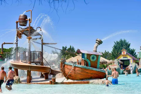 Scivolo e giostra acquatica rappresentante una barca con adulti e bambini che giocano in piscina in costume da bagno al parco Aquafollie a Caorle