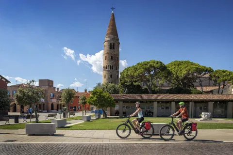 Due ciclisti attraversano il centro di Caorle. Sullo sfondo il caratteristico campanile del Duomo.