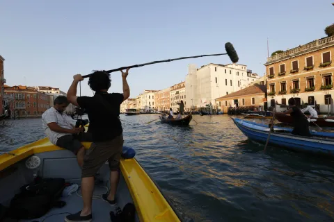 Due operatori della troupe dalla barca stanno riprendendo quattro imbarcazioni di vogatori in barca sul Canal Grande di Venezia. Sullo sfondo si vedono i palazzi veneziani affacciati sul canale e la fermata di un vaporetto con il battello in partenza. 