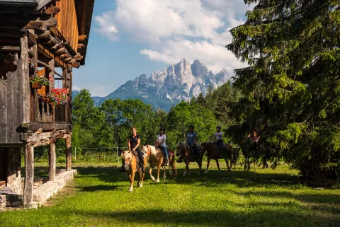 Alcune persone passeggiano a cavallo in uno spiazzo davanti ad una abitazione in Val di Zoldo