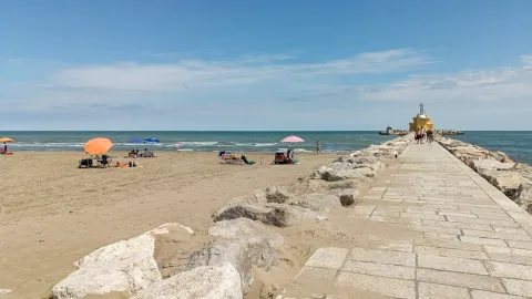 Spiaggia di Punta Sabbioni con bagnanti sdraiati su asciugamani sotto gli ombrelloni in una giornata di sole con passerella e faro giallo a Cavallino Treporti