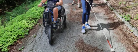 Una persona in sedia a rotelle e una persona cieca con bastone camminano lungo il tratto asfaltato del Sentiero della Sensibilità